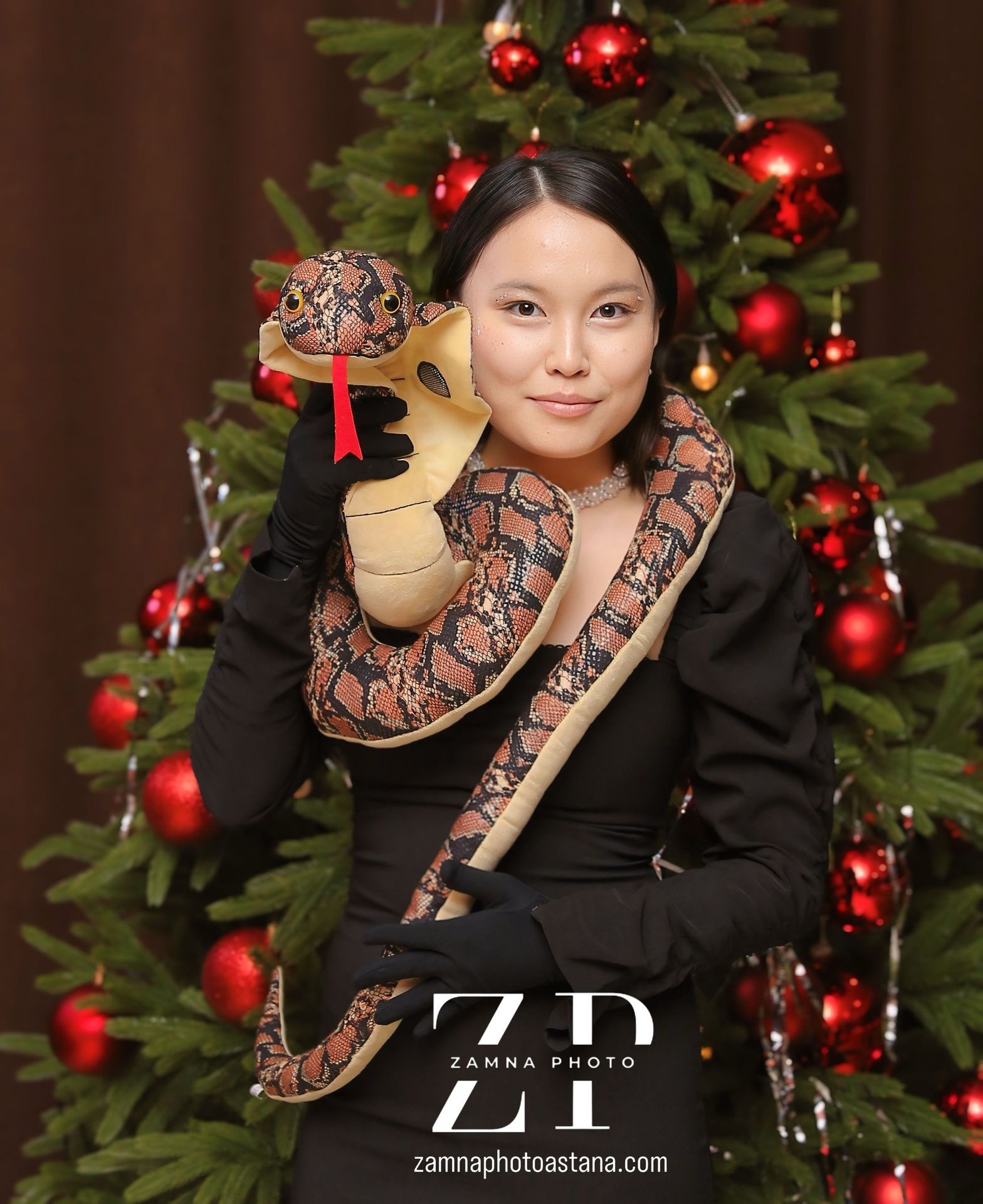 Person in black dress holding a stuffed snake toy, standing in front of a decorated Christmas tree.