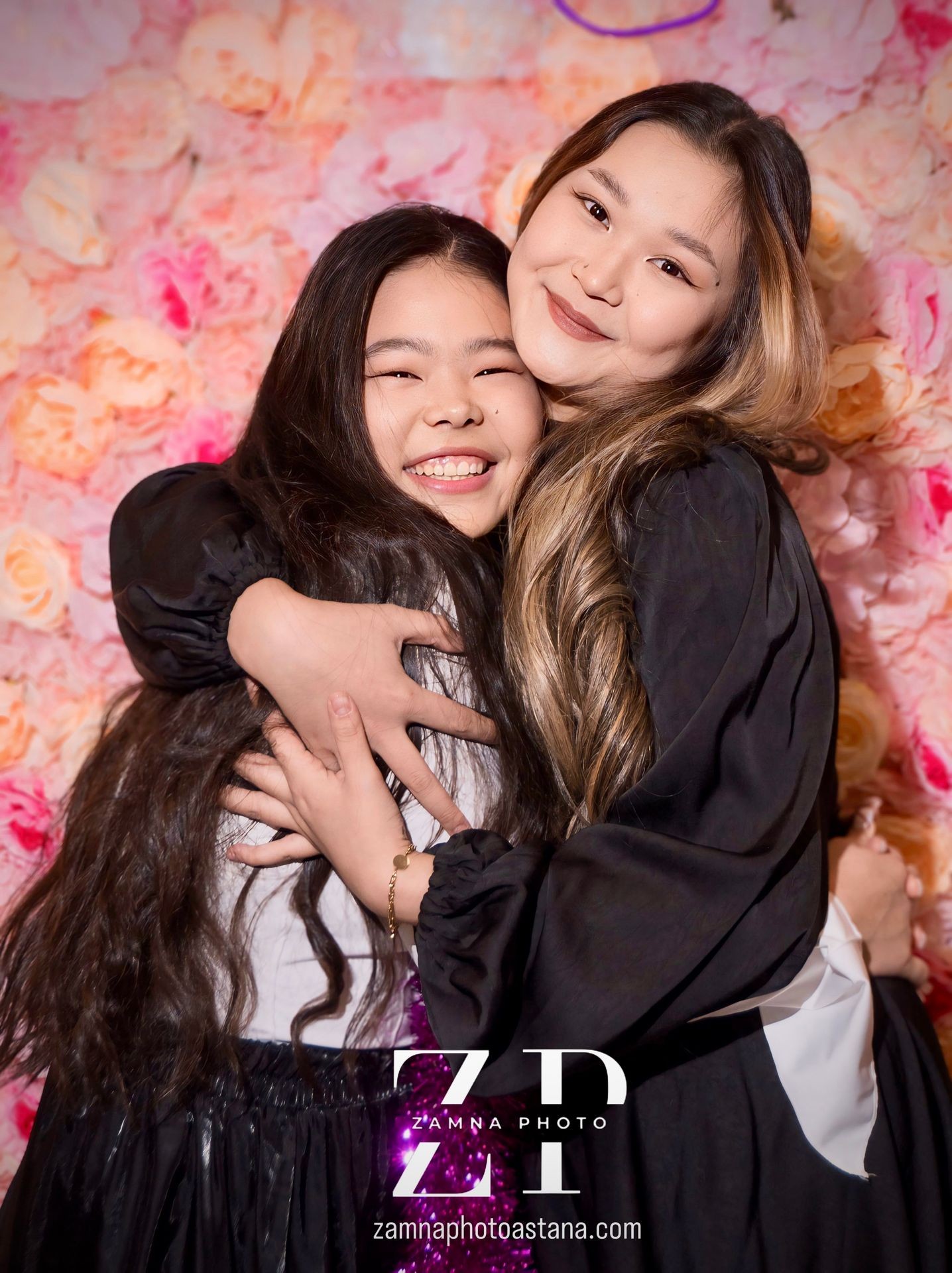 Two people hugging and smiling in front of a pink floral backdrop.