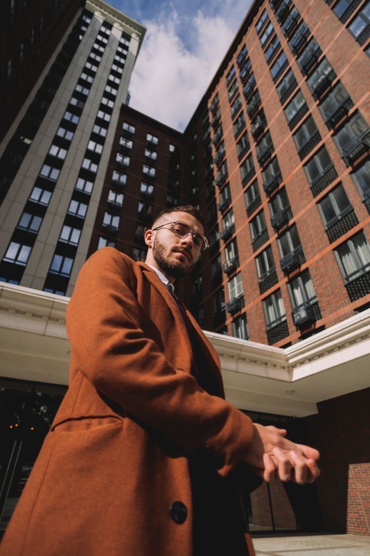 Man in brown coat standing in front of tall brick buildings under a blue sky.