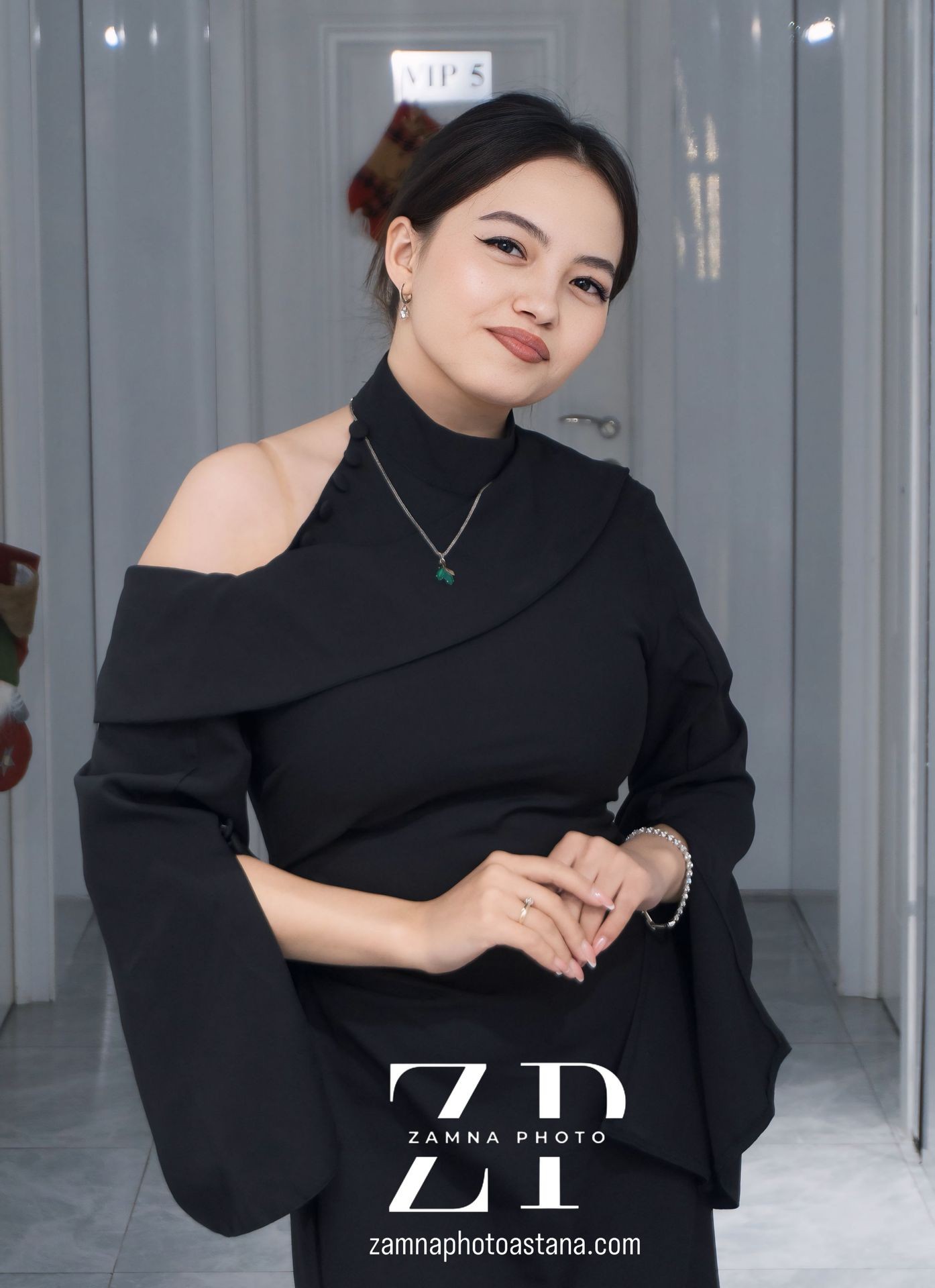 Woman in a black dress posing indoors with hands clasped and smiling.