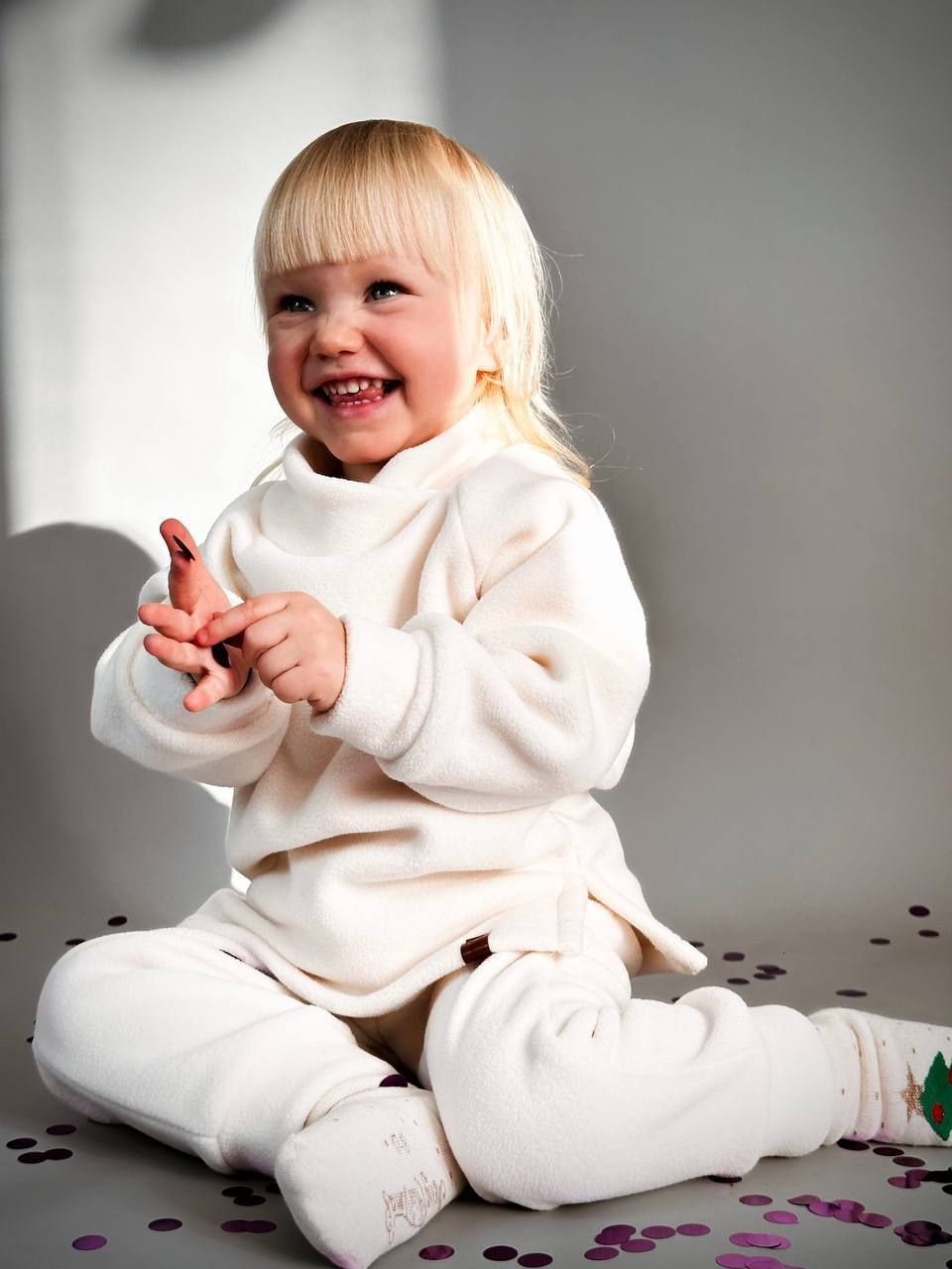 Smiling toddler in a white outfit sitting on the floor surrounded by confetti.