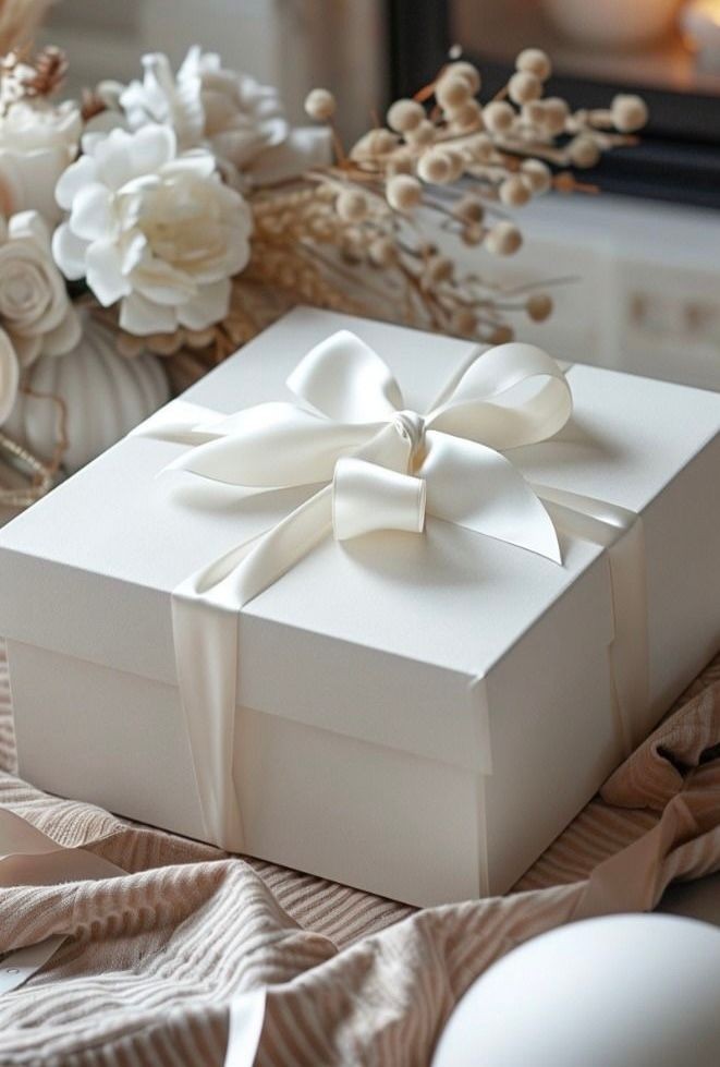 Elegant white gift box with satin ribbon surrounded by flowers and decor on a textured surface.