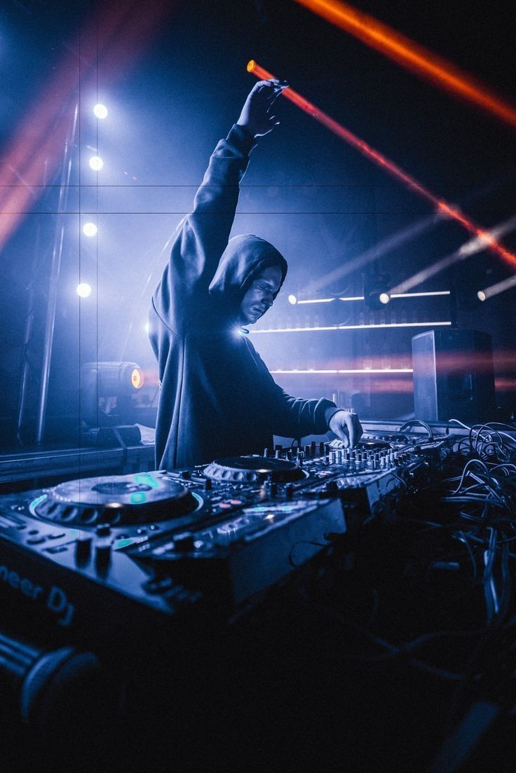 DJ wearing a hoodie performing on stage with mixing equipment and colorful lights in the background.