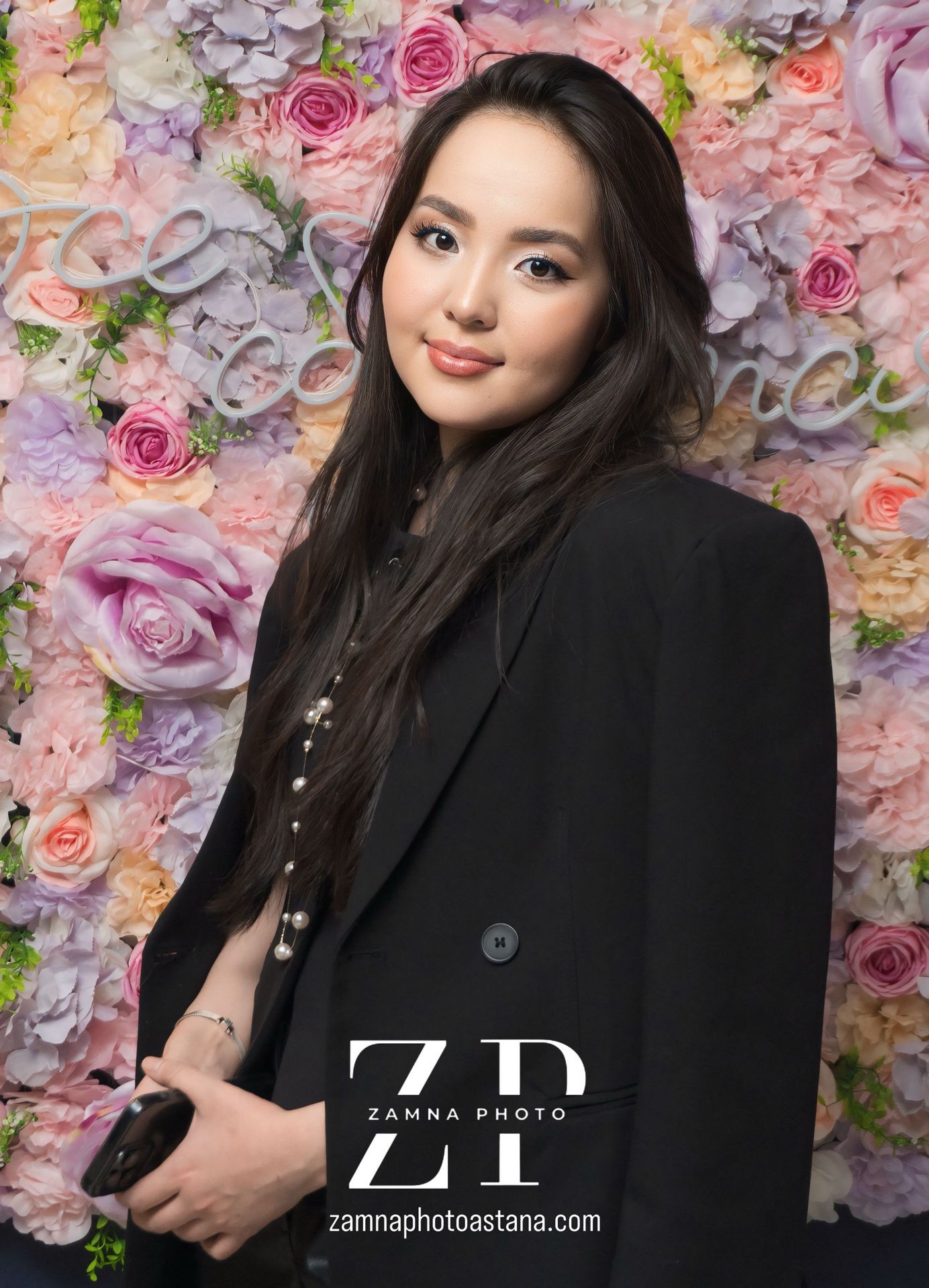 Woman in black coat standing in front of a flower wall holding a phone.