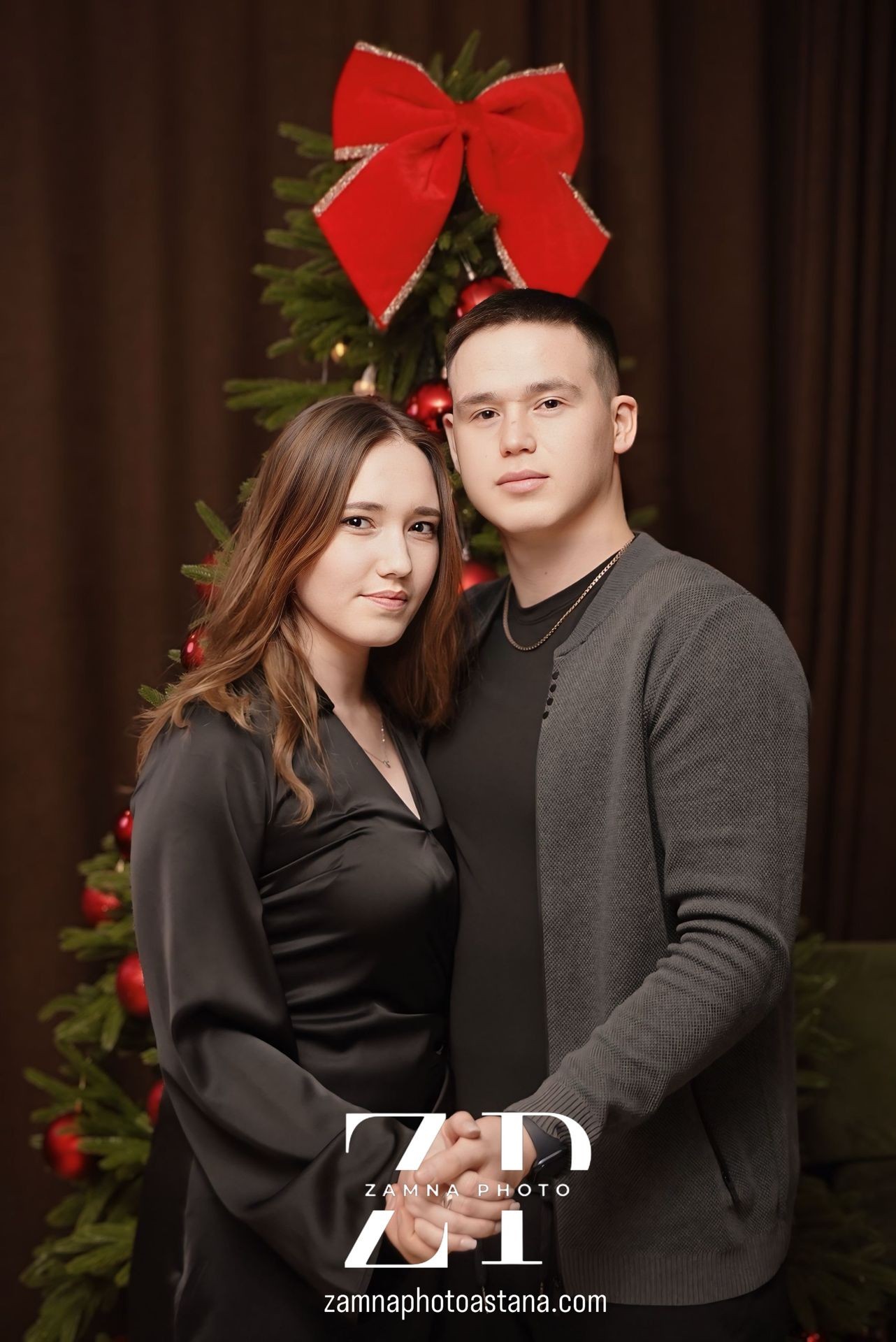 Couple posing in front of a Christmas tree with red decorations and a large red bow.