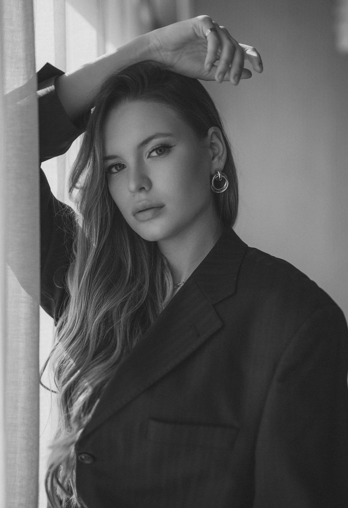 Black and white portrait of a woman in a blazer with long hair, standing by a window.