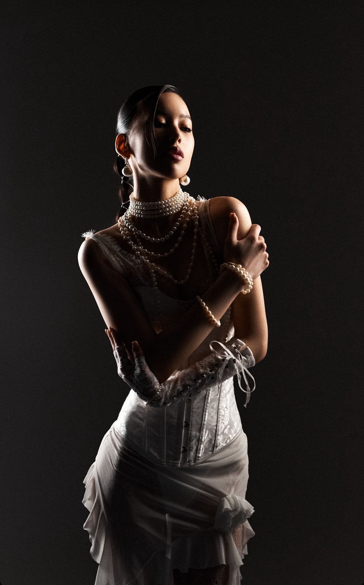 Woman in elegant white dress with pearl jewelry poses against a dark background.